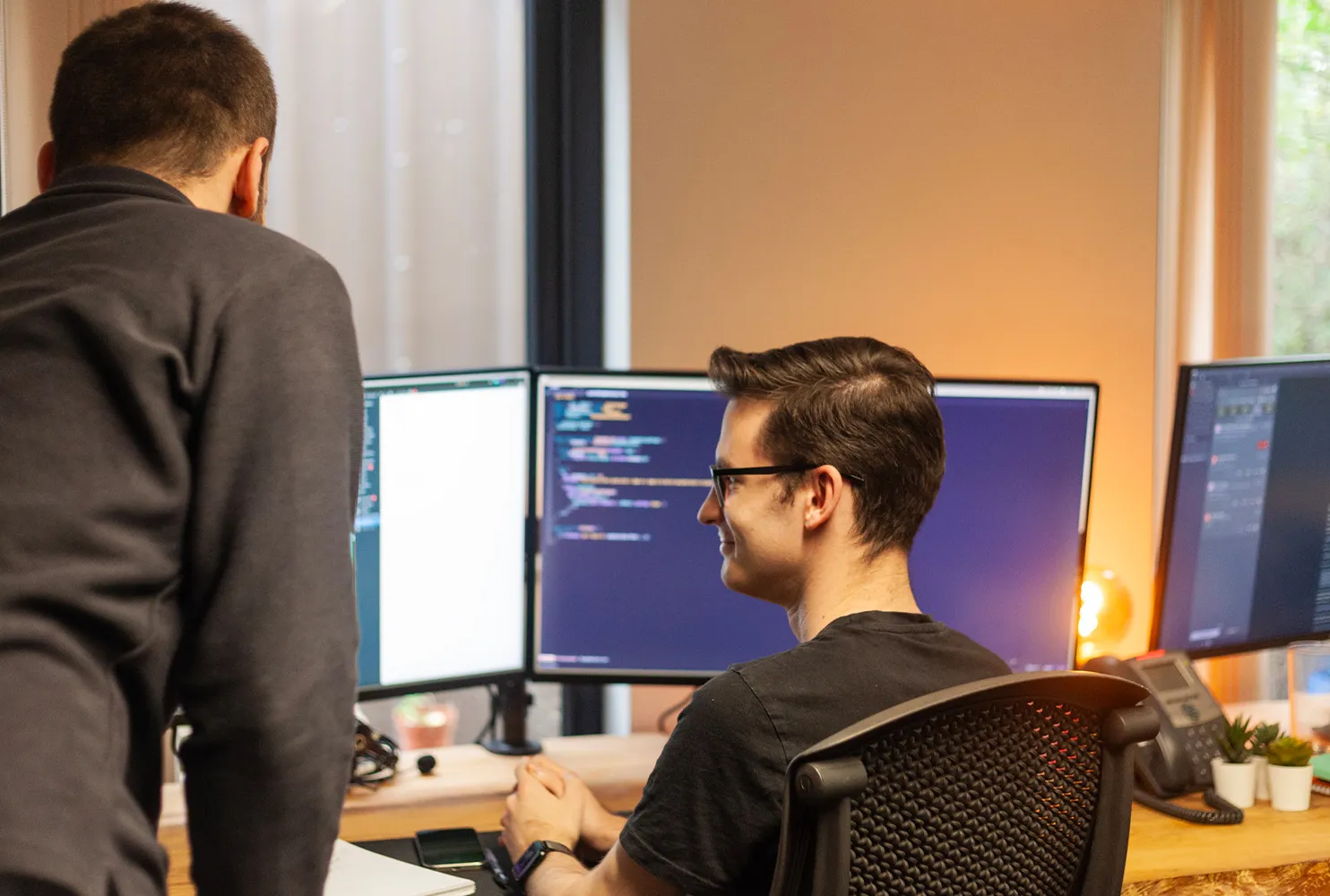 Two people in an office setup with multiple monitors displaying code