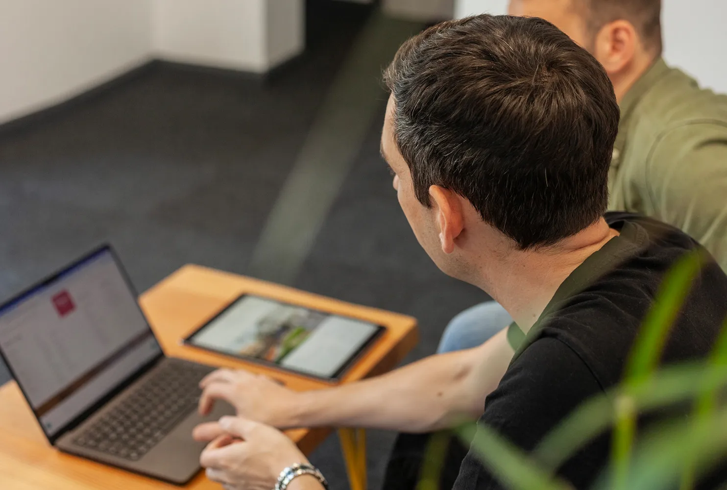 Two men sitting at a wooden table focus on a laptop and tablet