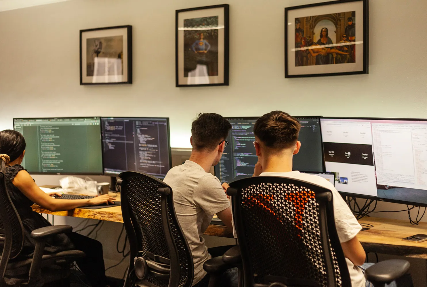 Three people working at a wooden desk in an office focusing on coding on large monitors