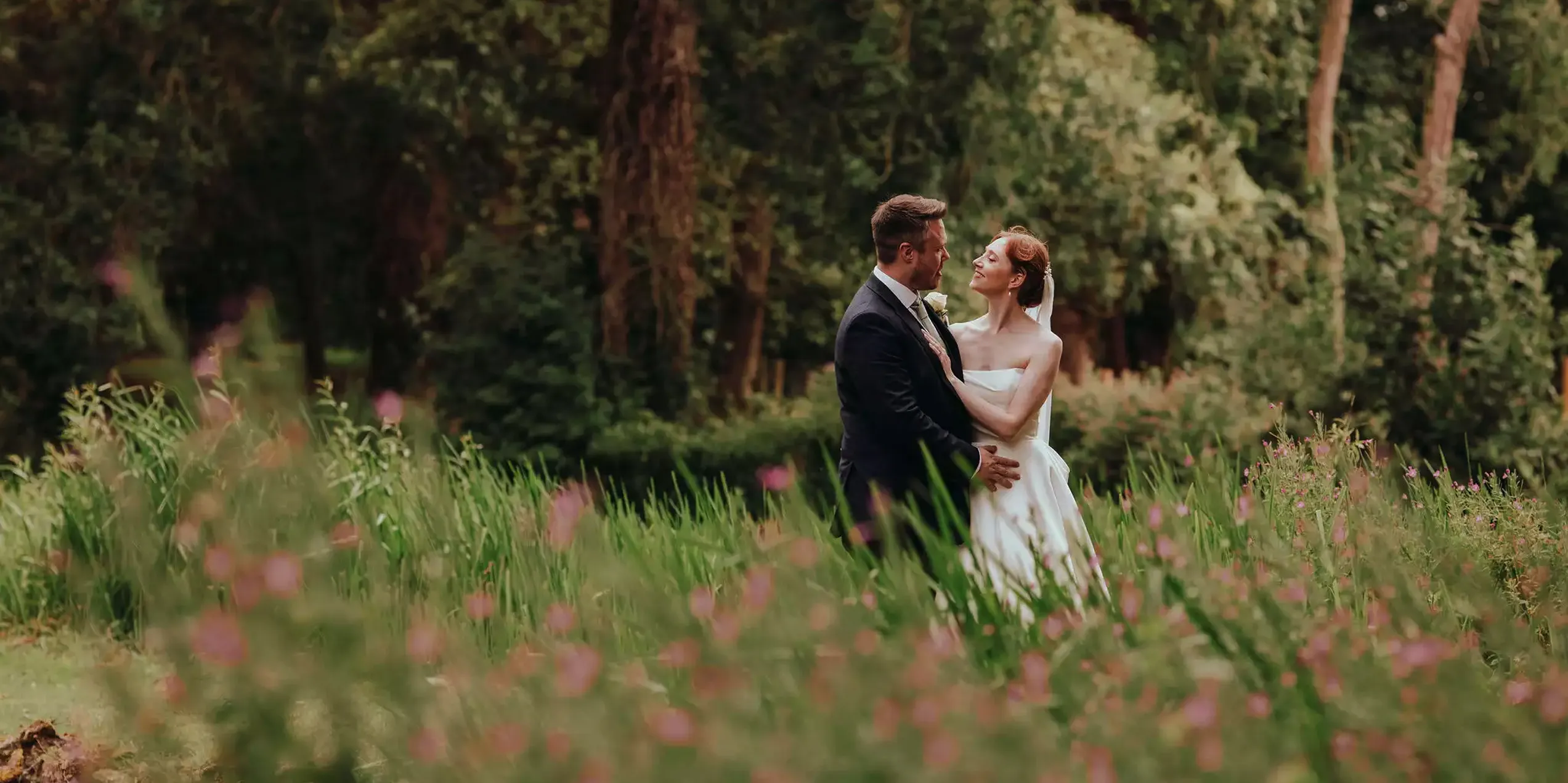 A bride and groom embrace in a lush garden, surrounded by greenery and pink wildflowers.