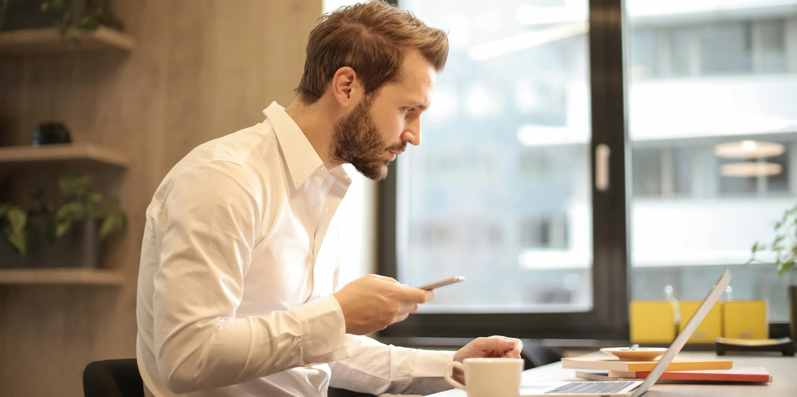 A man sat at a desk holding a phone whilst using a laptop