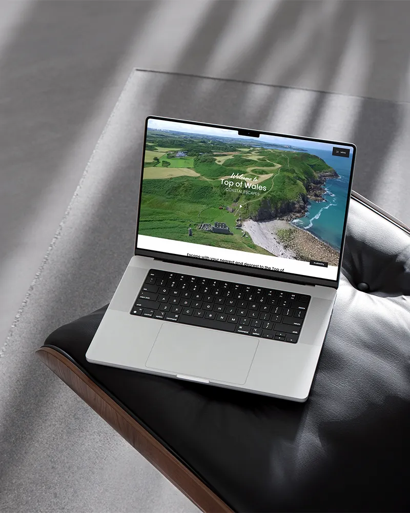 A MacBook on a black leather seat showing the Top of Wales website homepage