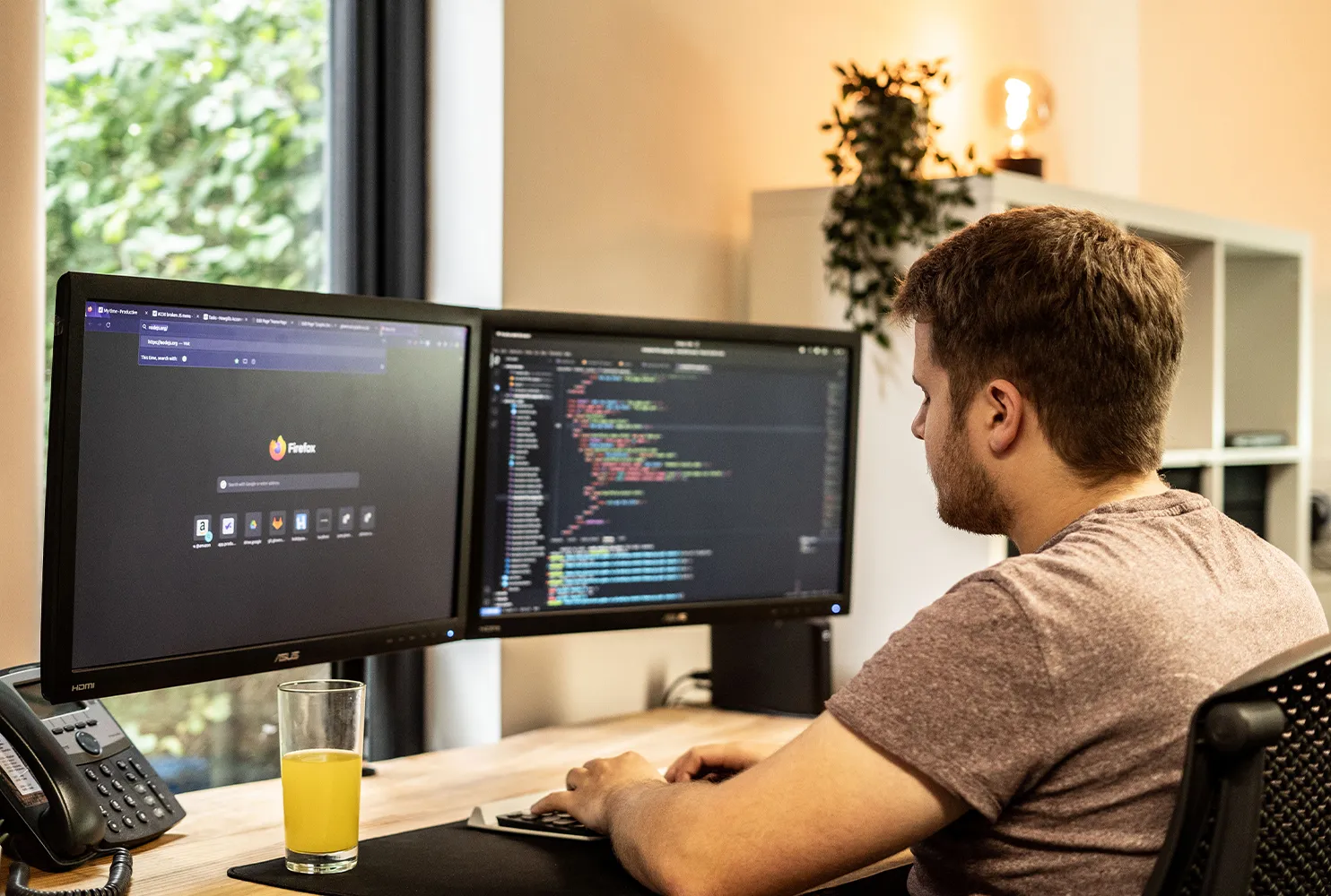 A man sitting at a wooden desk looking at computer screens showing lines of code