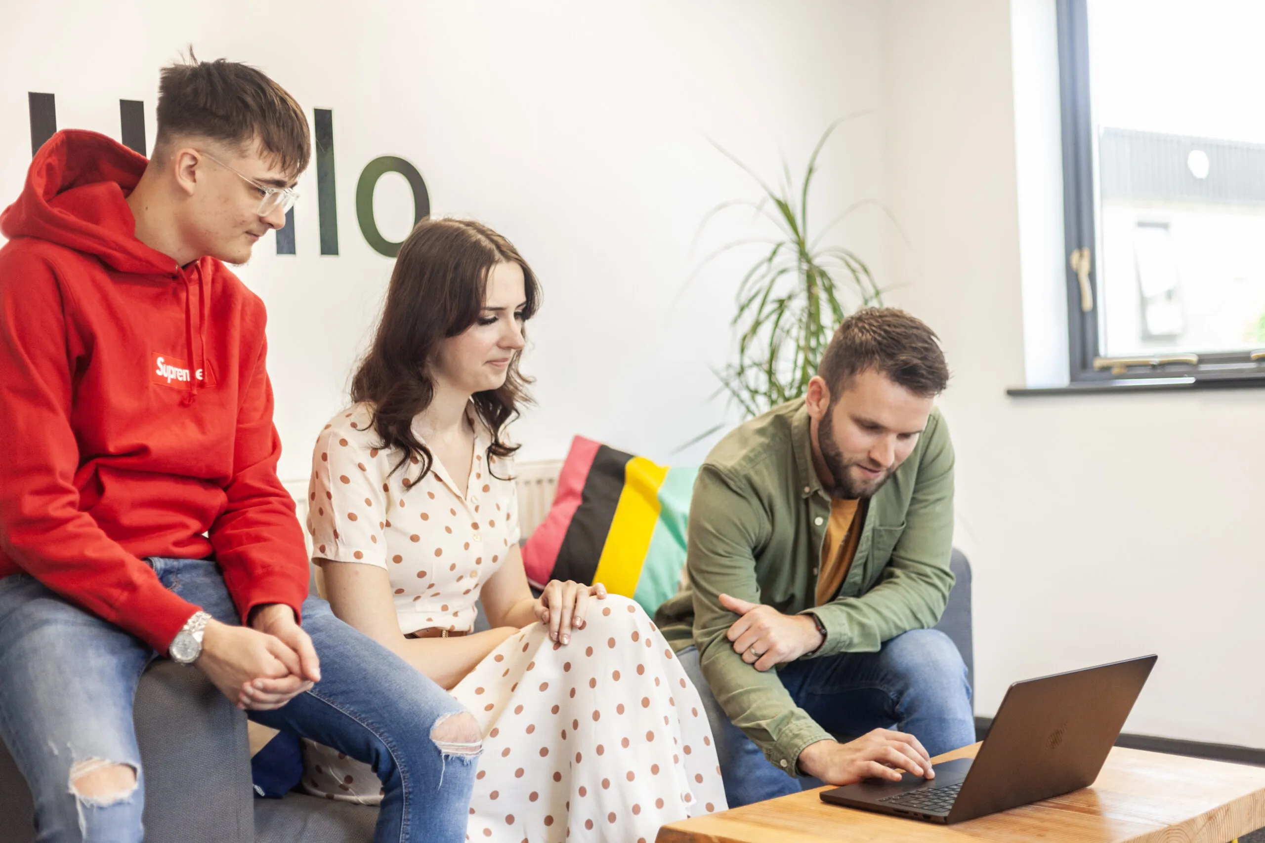 Two men and a woman sitting on a grey sofa looking down at a MacBook on a coffee table