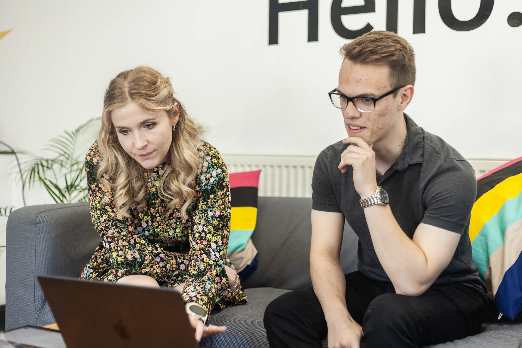 A man and a woman sitting on a grey sofa looking at a MacBook