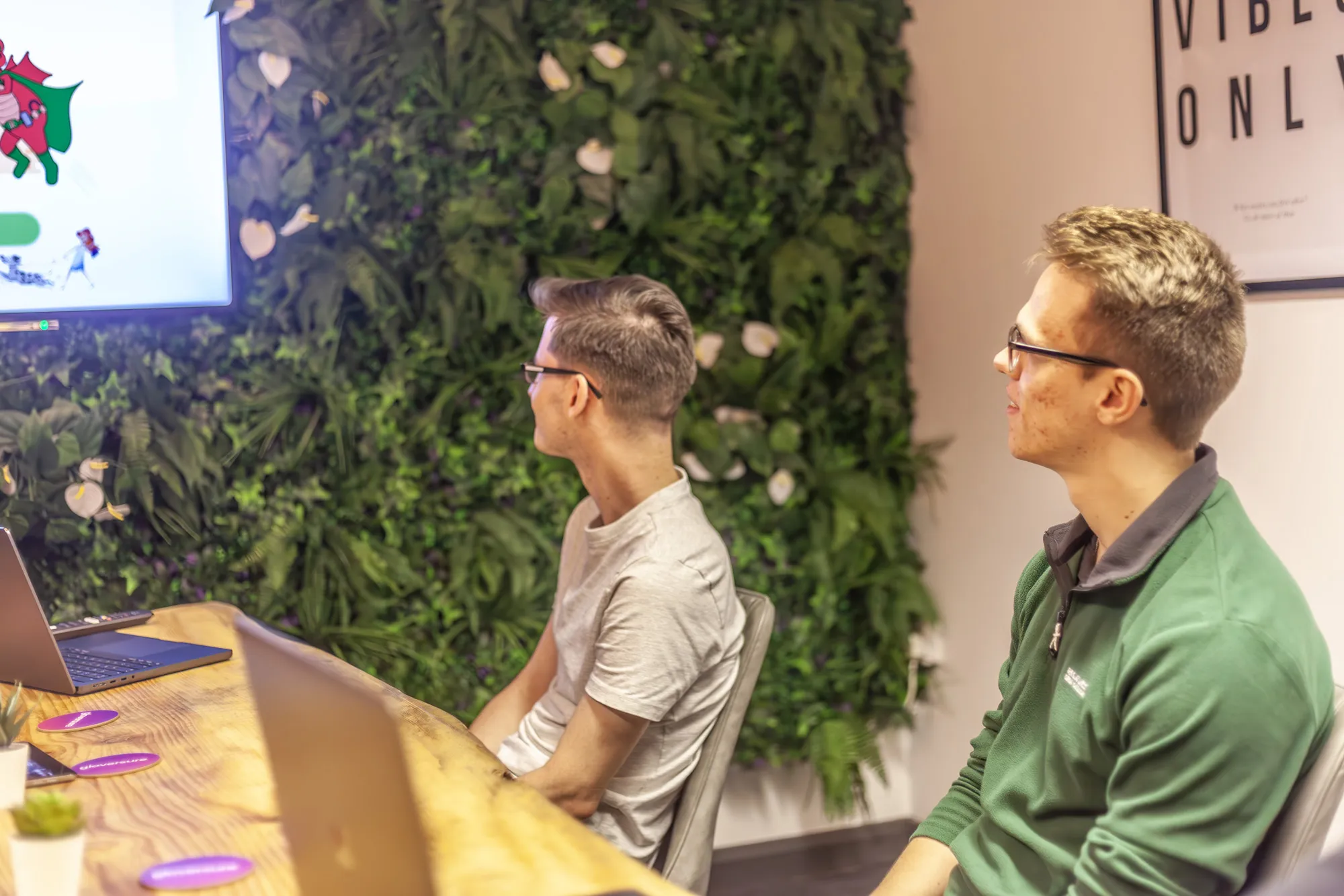 Two men sitting at a wooden table looking towards a wall mounted TV screen