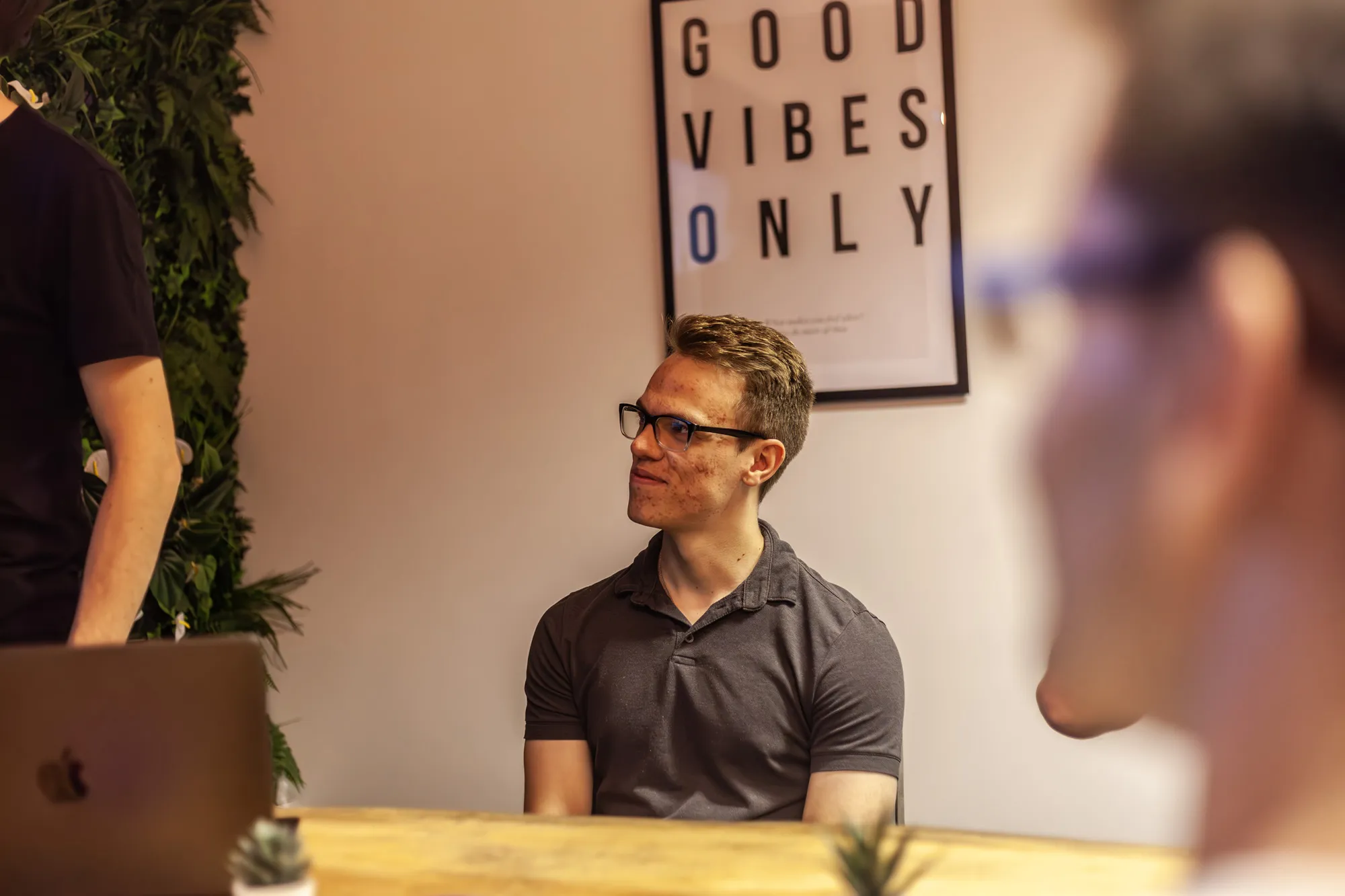 A man sitting at a wooden desk with a MacBook looking on