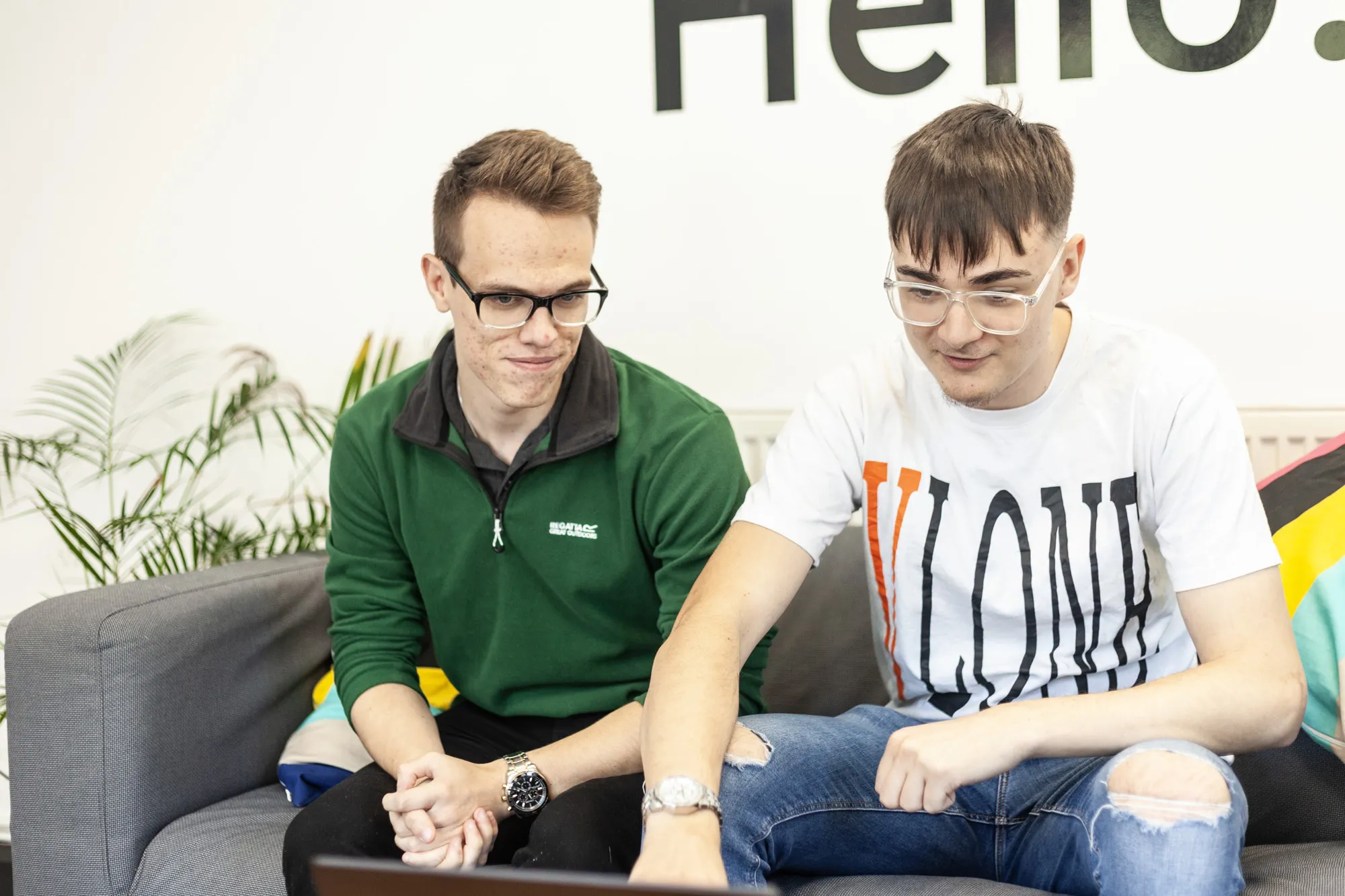 Two men sat on a grey sofa looking down at a MacBook