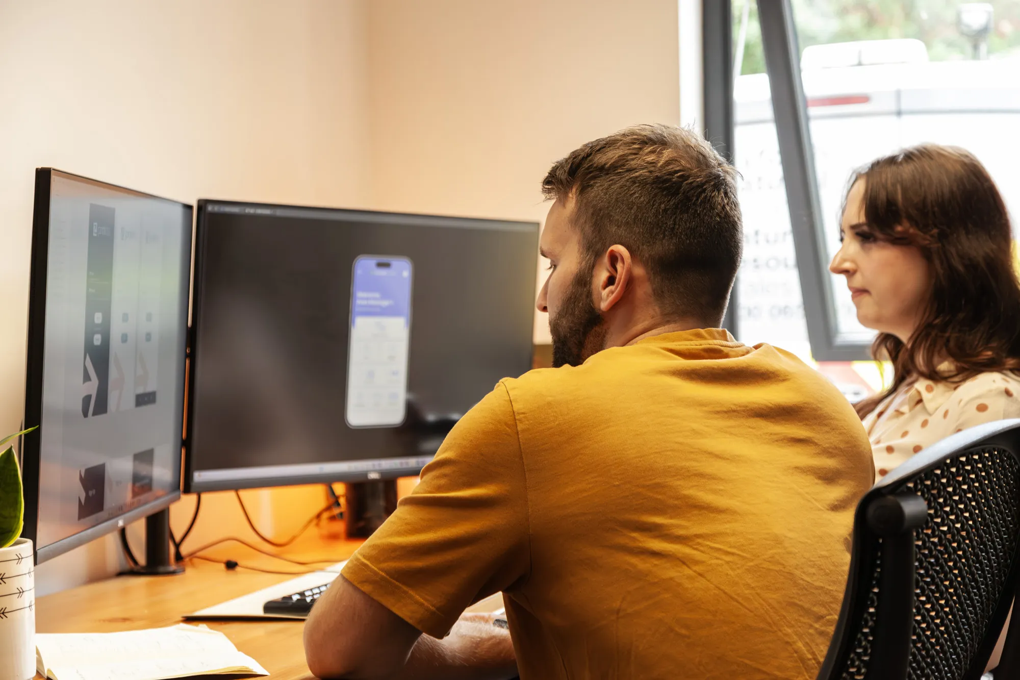 A man and a woman sat at a desk looking at a computer screen with mobile app prototypes on screen