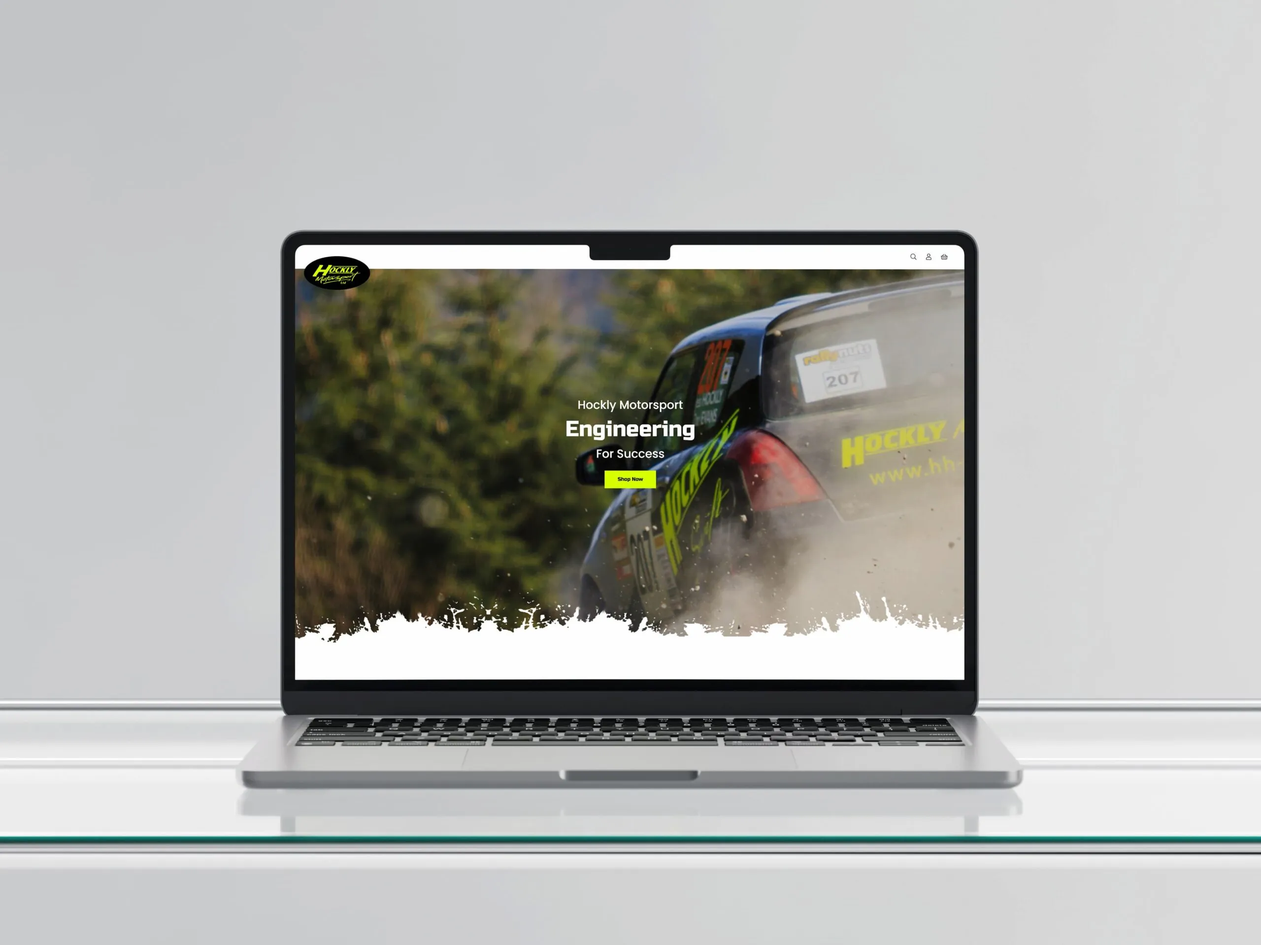 A MacBook on a glass table showing the Harry Hockly website homepage onscreen