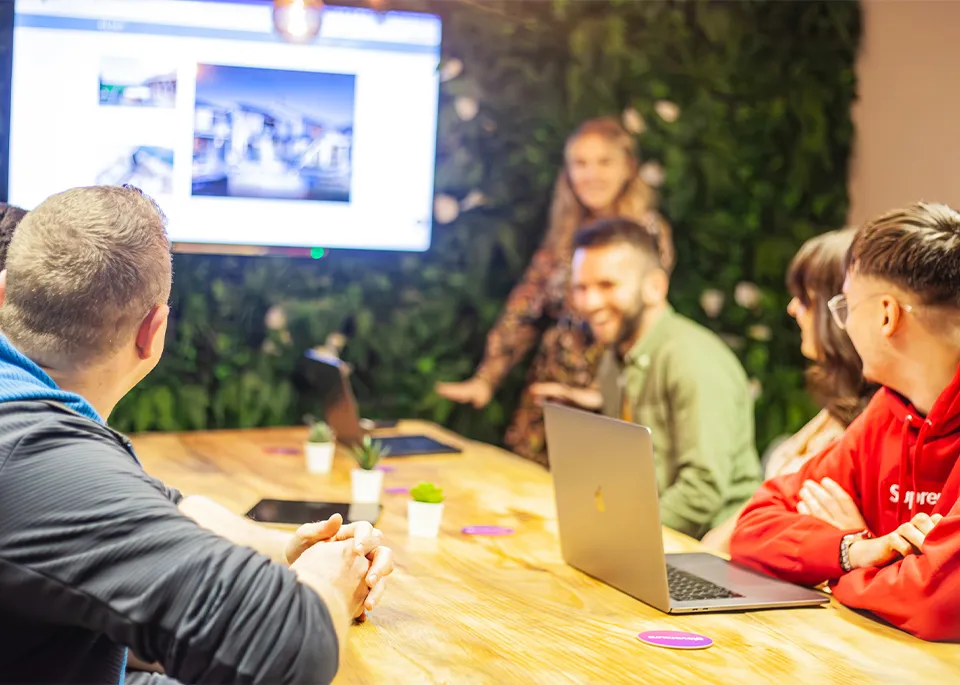 A group of people sat around a wooden table with laptops laughing