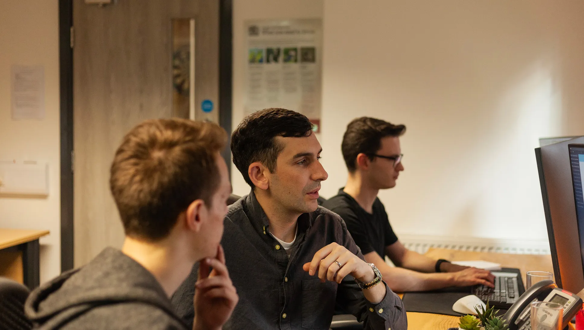 Three men sitting at a wooden desk looking at computer screens