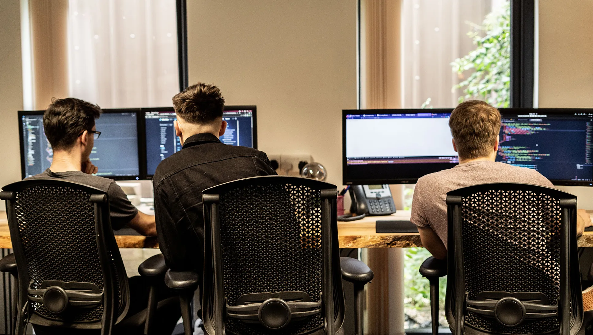 Three men sat at a long wooden desk looking at computer screen with lines of code