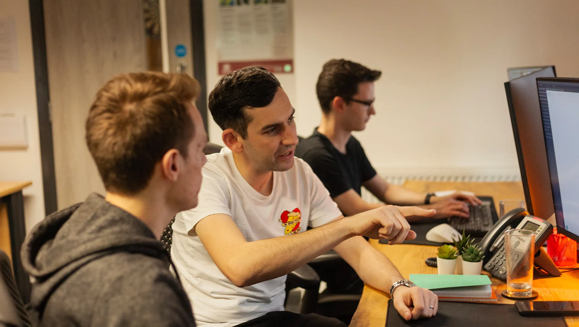 Three men sat at a wooden desk looking on