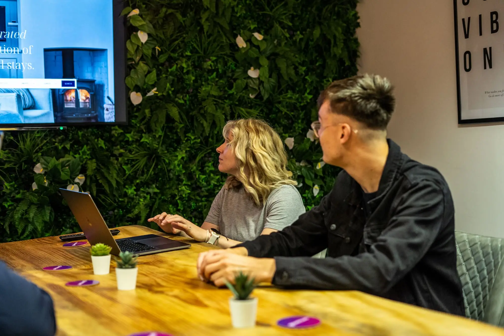 A man and a woman sitting at a wooden table with a laptop looking on