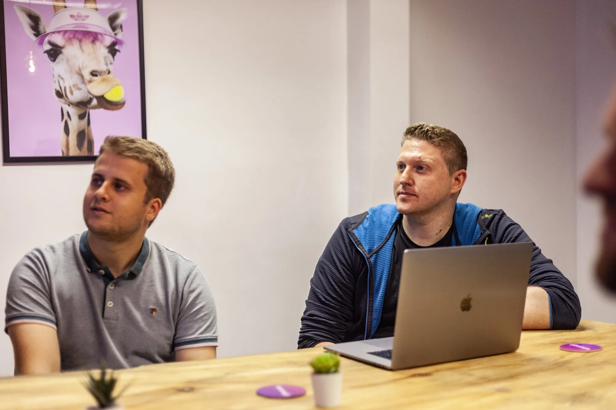 Two men sat at a wooden table with a MacBook