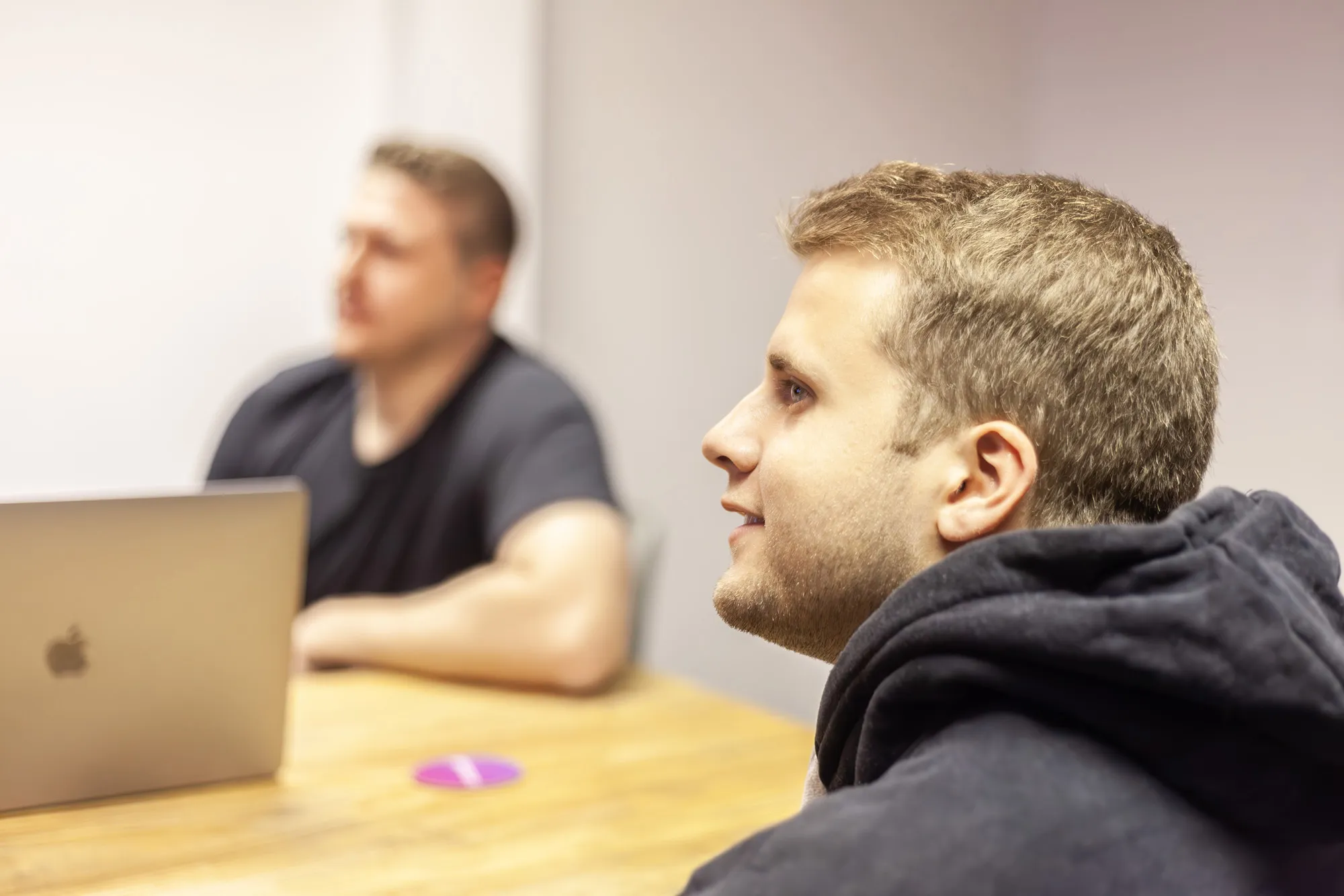 Two men at a wooden table with a MacBook looking on