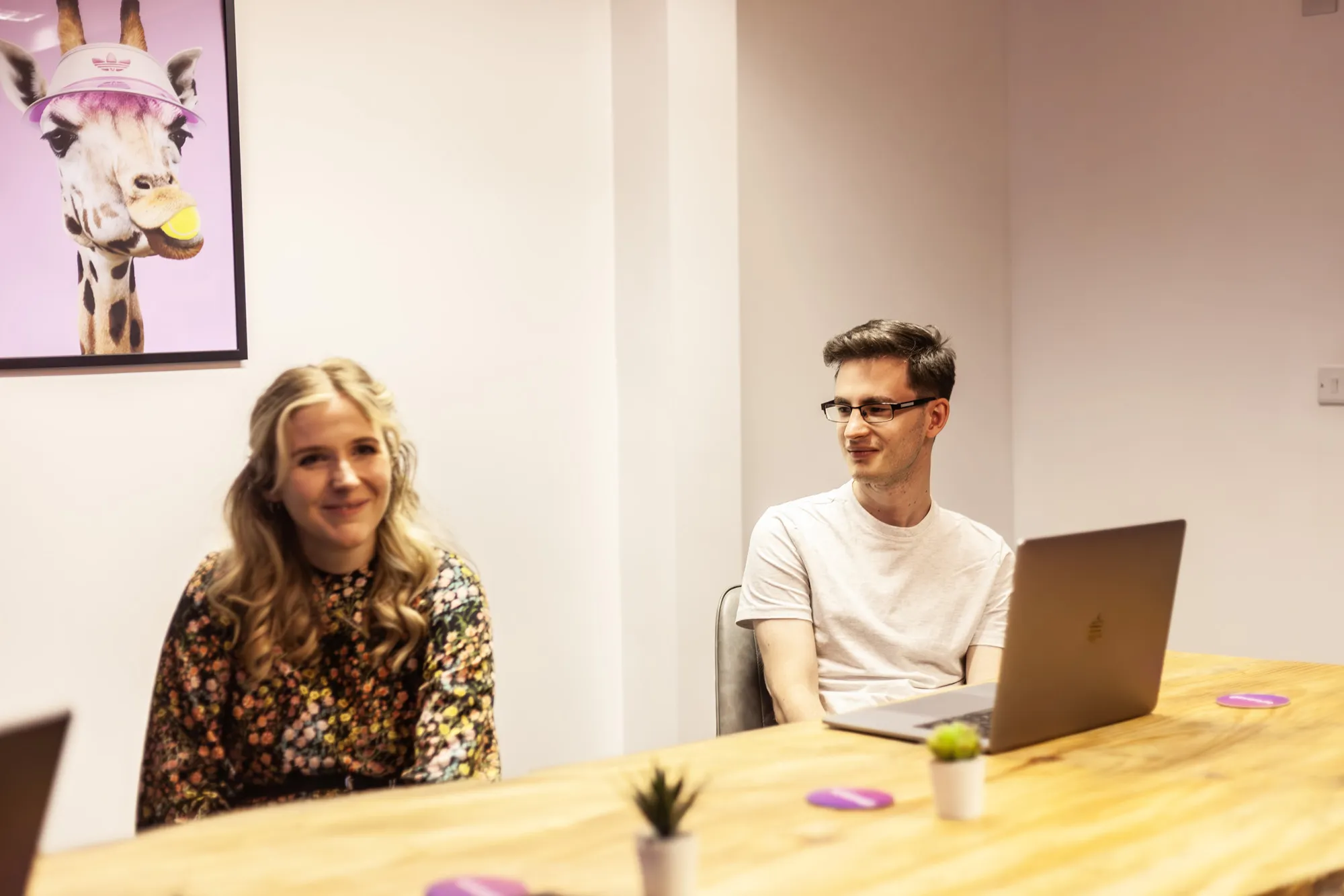 A man and a woman sat at a wooden table with a MacBook