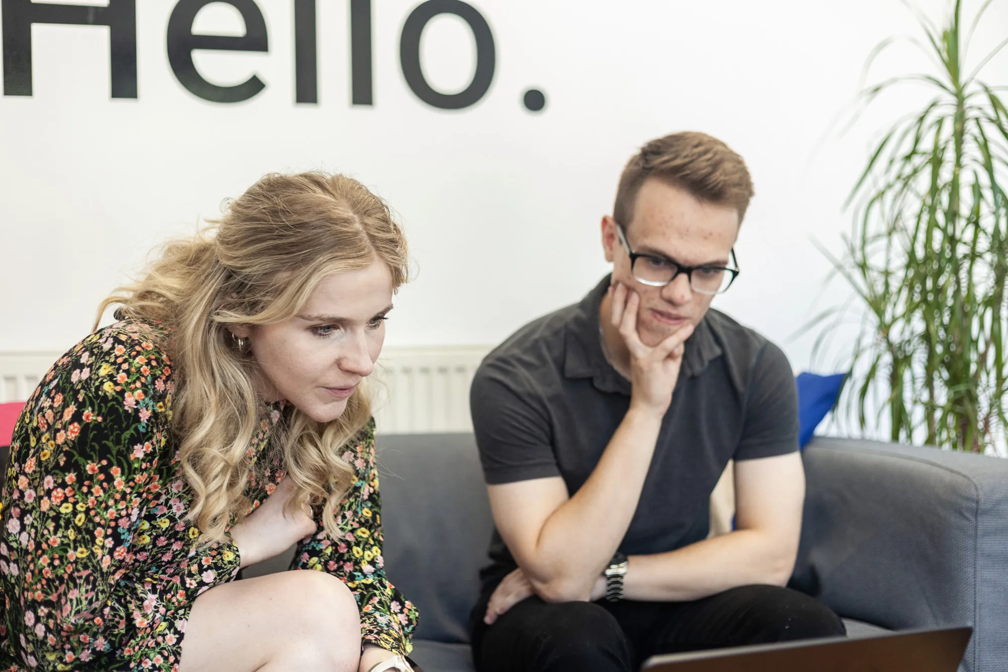A man and a woman sat on a grey sofa looking down at a MacBook
