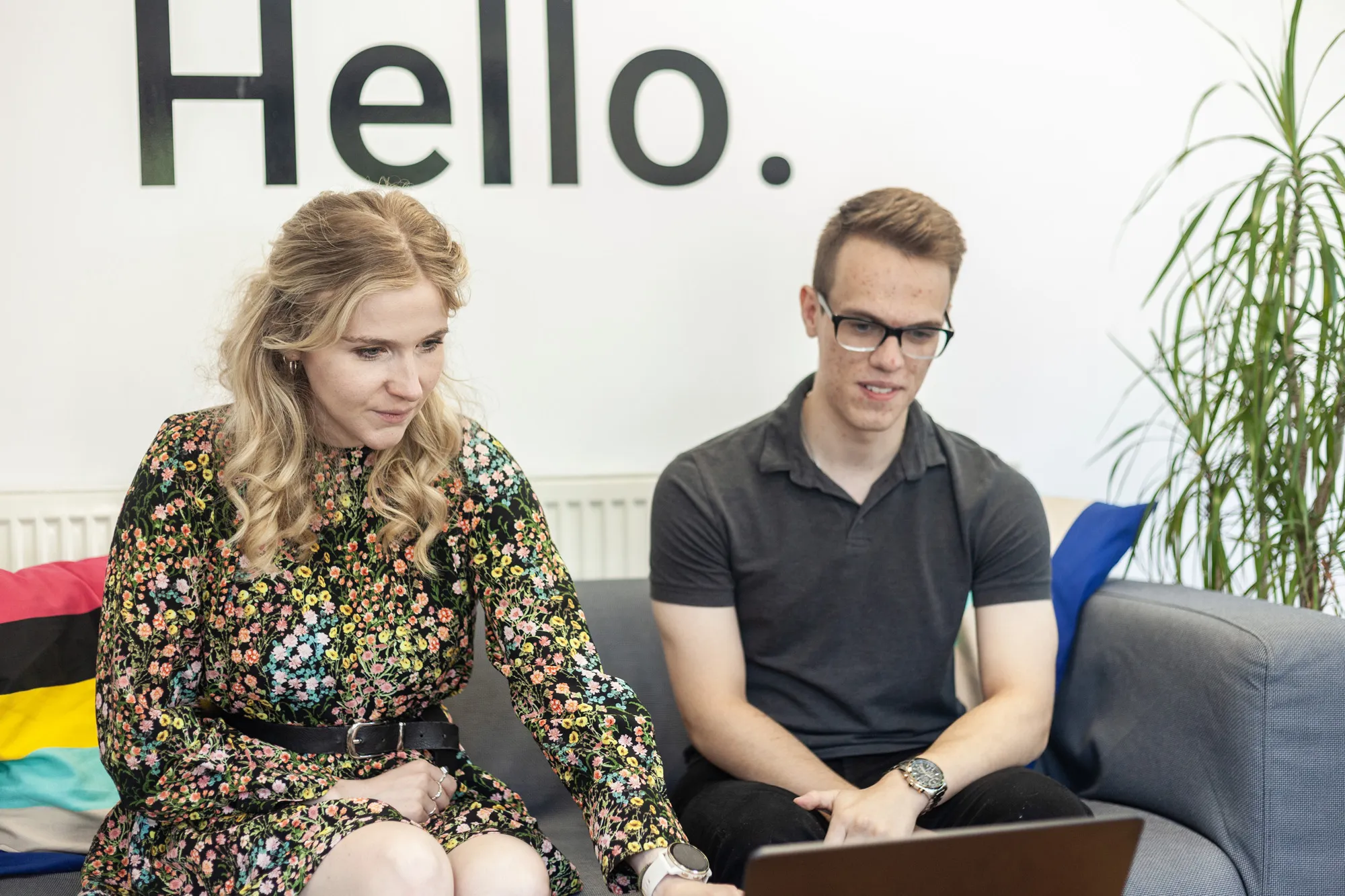 A man and a woman sat on a grey sofa looking down at a MacBook 