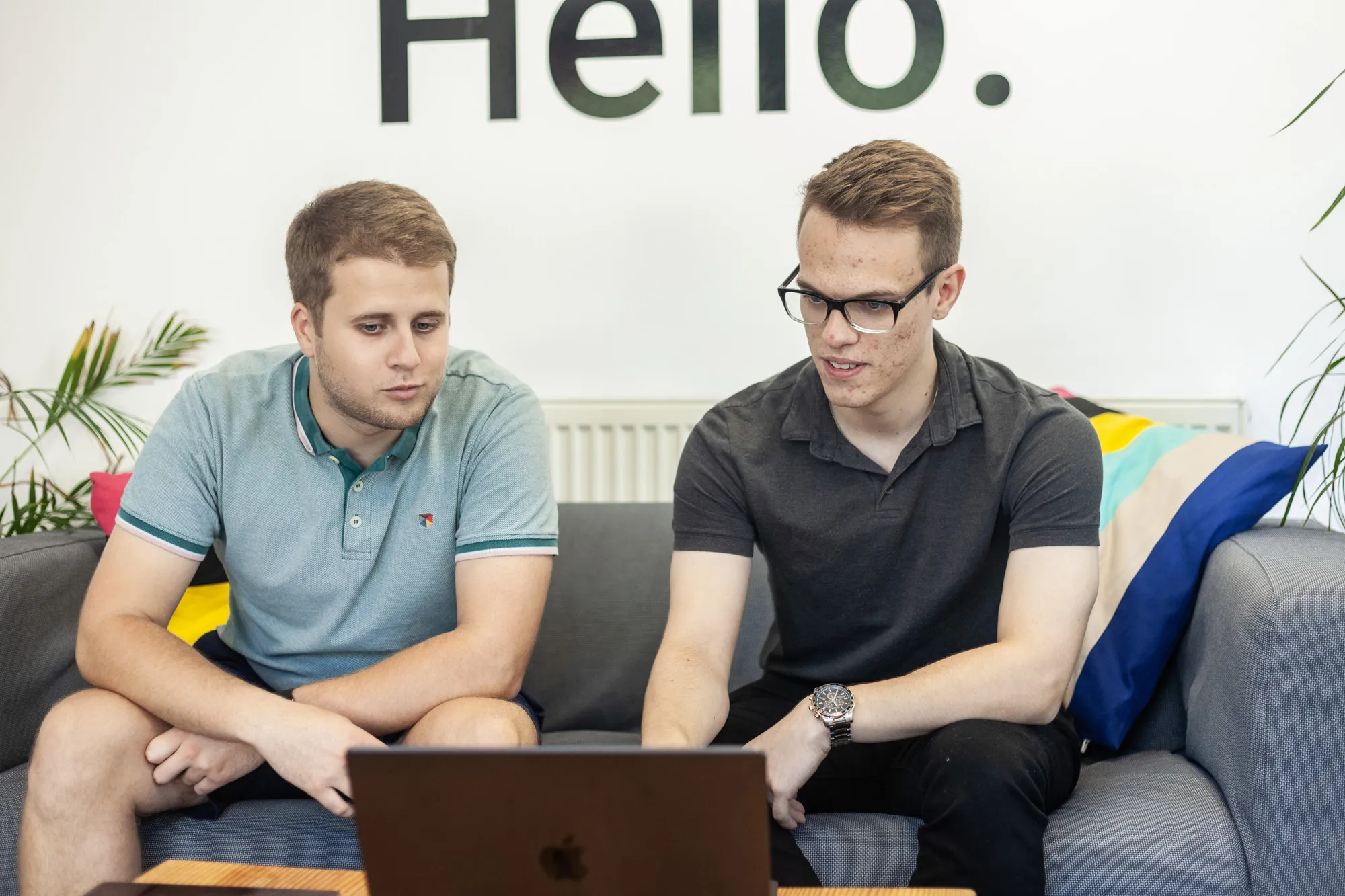 Two men sat on a grey sofa looking at a MacBook