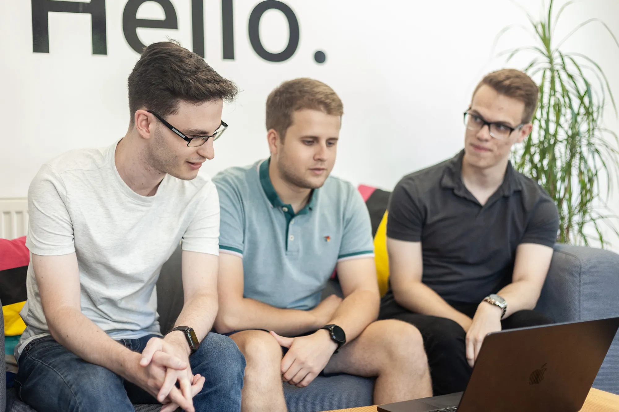 Three men sat on a grey sofa looking down at a MacBook