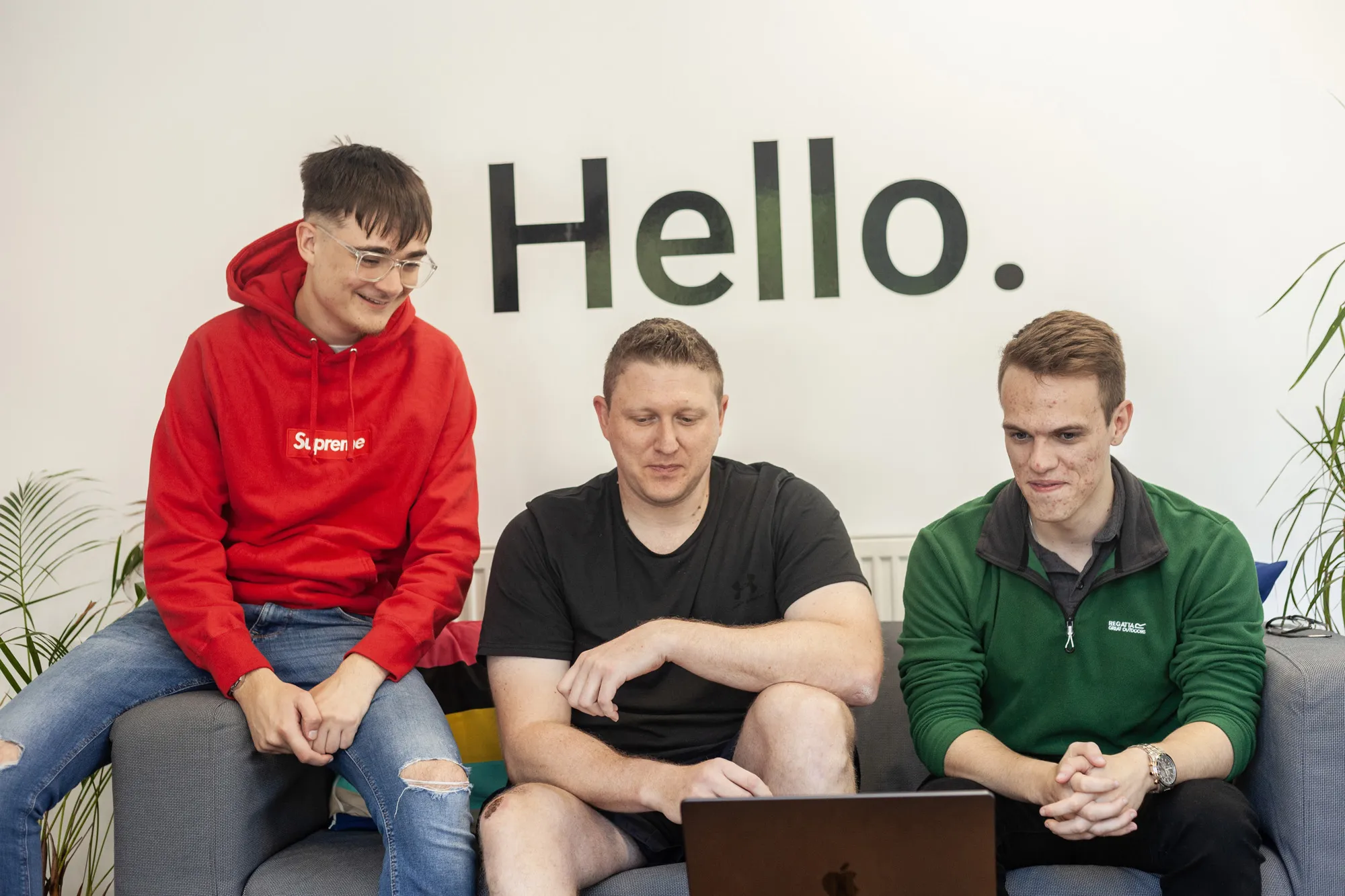 Three men sat on a grey sofa looking down at a MacBook