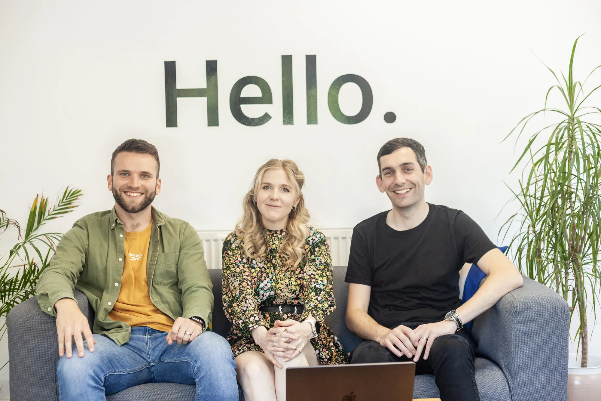 Two men and a woman sat on a grey sofa smiling