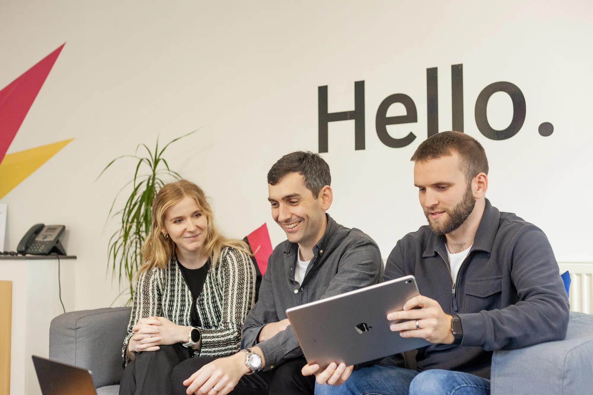 Two men and a woman sitting on a grey sofa looking at an iPad