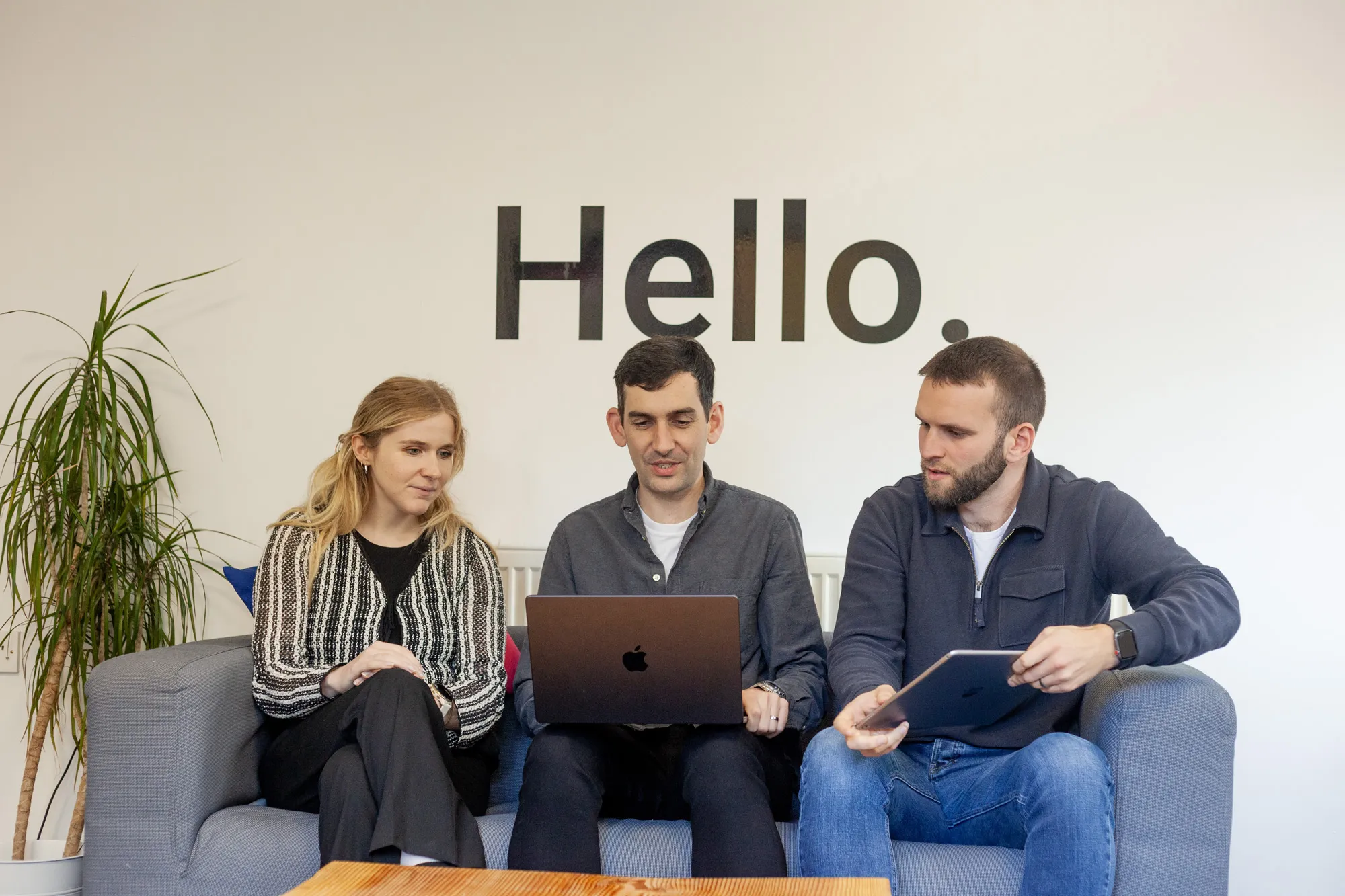 Two men and a woman sitting on a grey sofa looking at an iPad