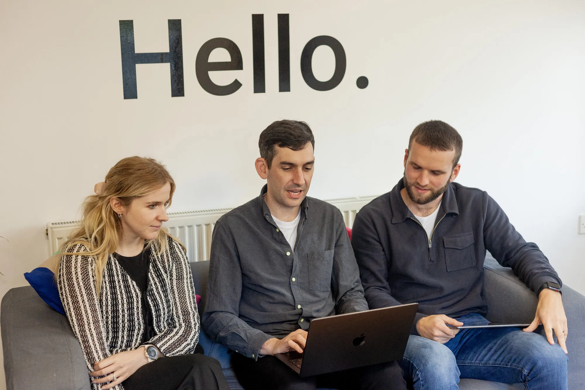 Two men and a woman sat on a grey sofa looking at a MacBook