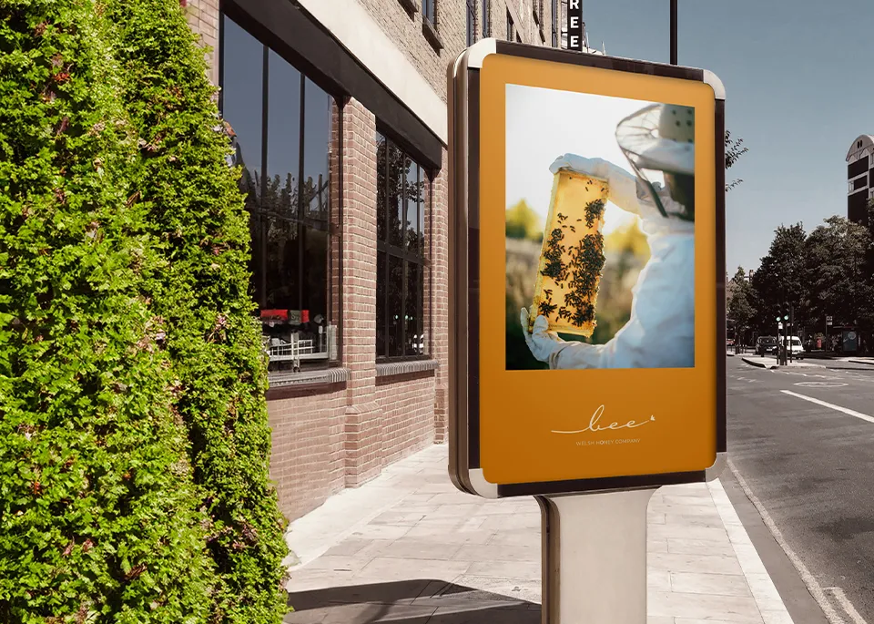 A billboard in a street showing the Bee Welsh Honey branding with the image of a beekeeper holding up a tray full of honey and bees