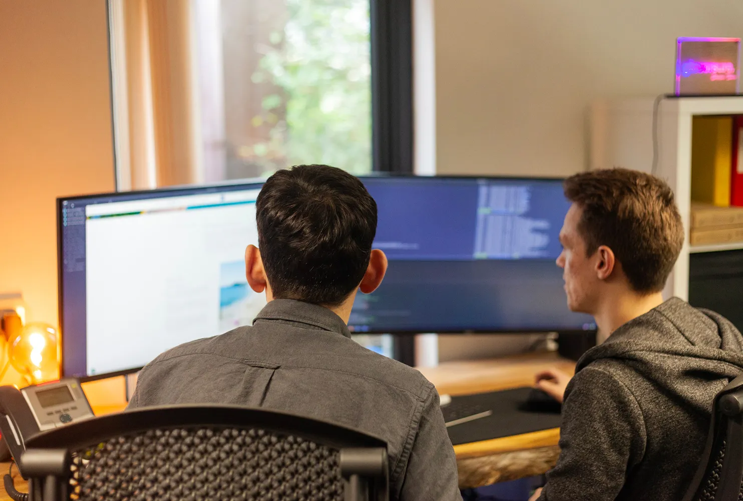 Two men sat at a wooden desk looking at computer screens with lines of code