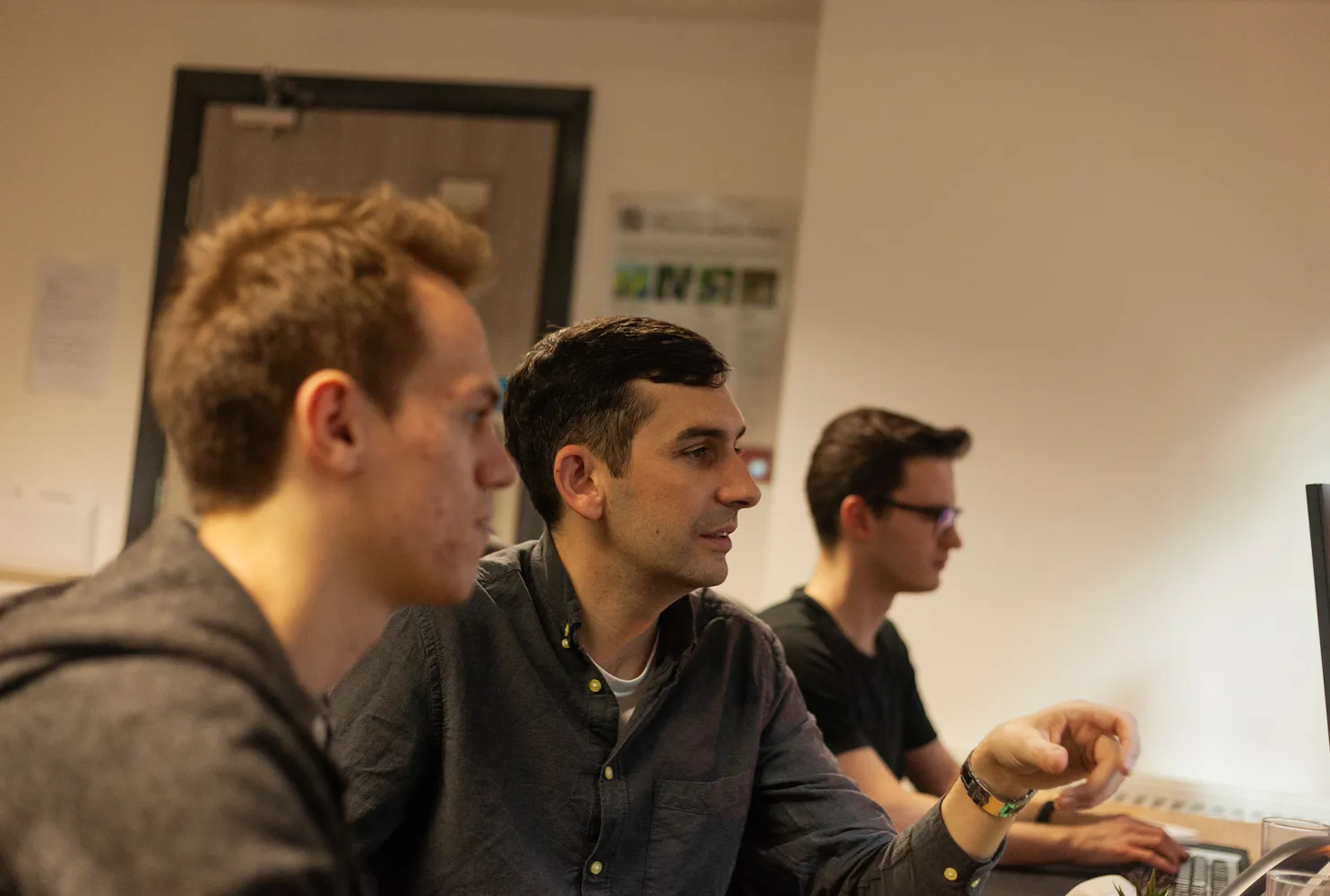 Three men sitting at a wooden desk looking at computer screens