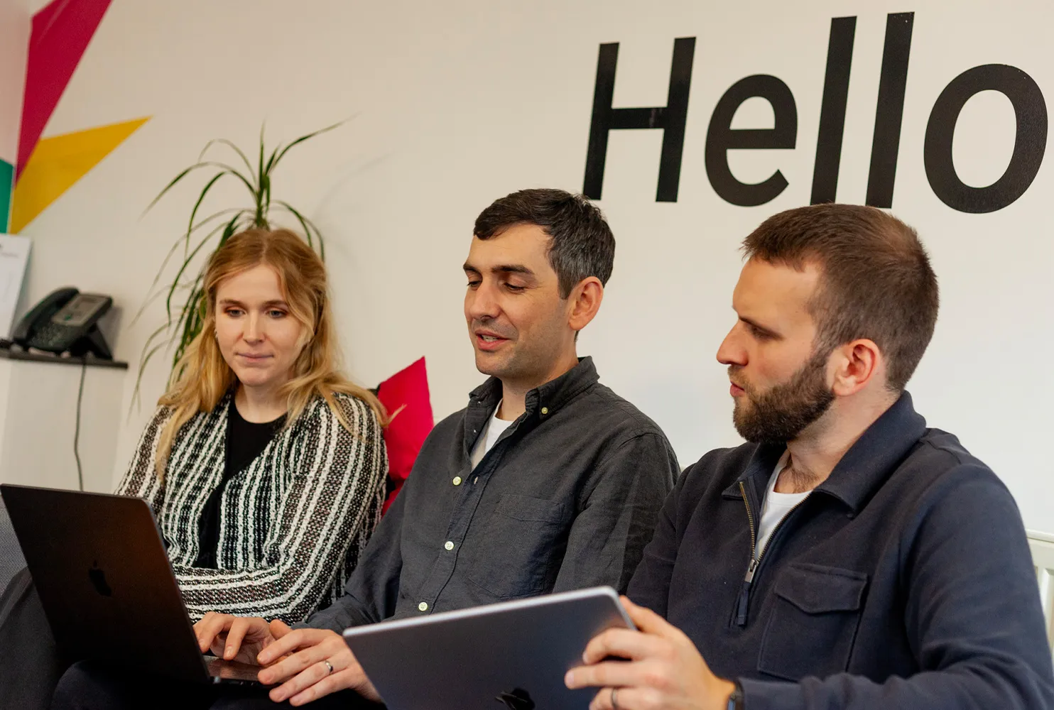 Two men and a woman sitting on a grey sofa looking at an iPad and a MacBook