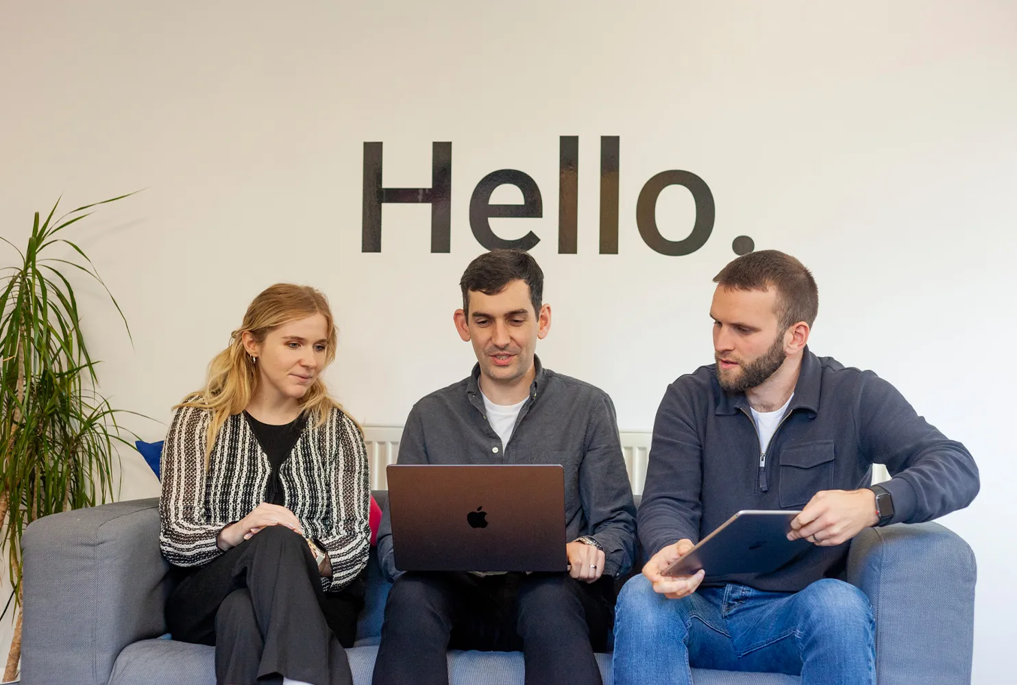 Two men and a woman sitting on a grey sofa looking at a laptop
