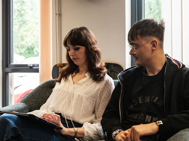 A woman in a white blouse sitting with an iPad next to a man in glasses and a dark jacket