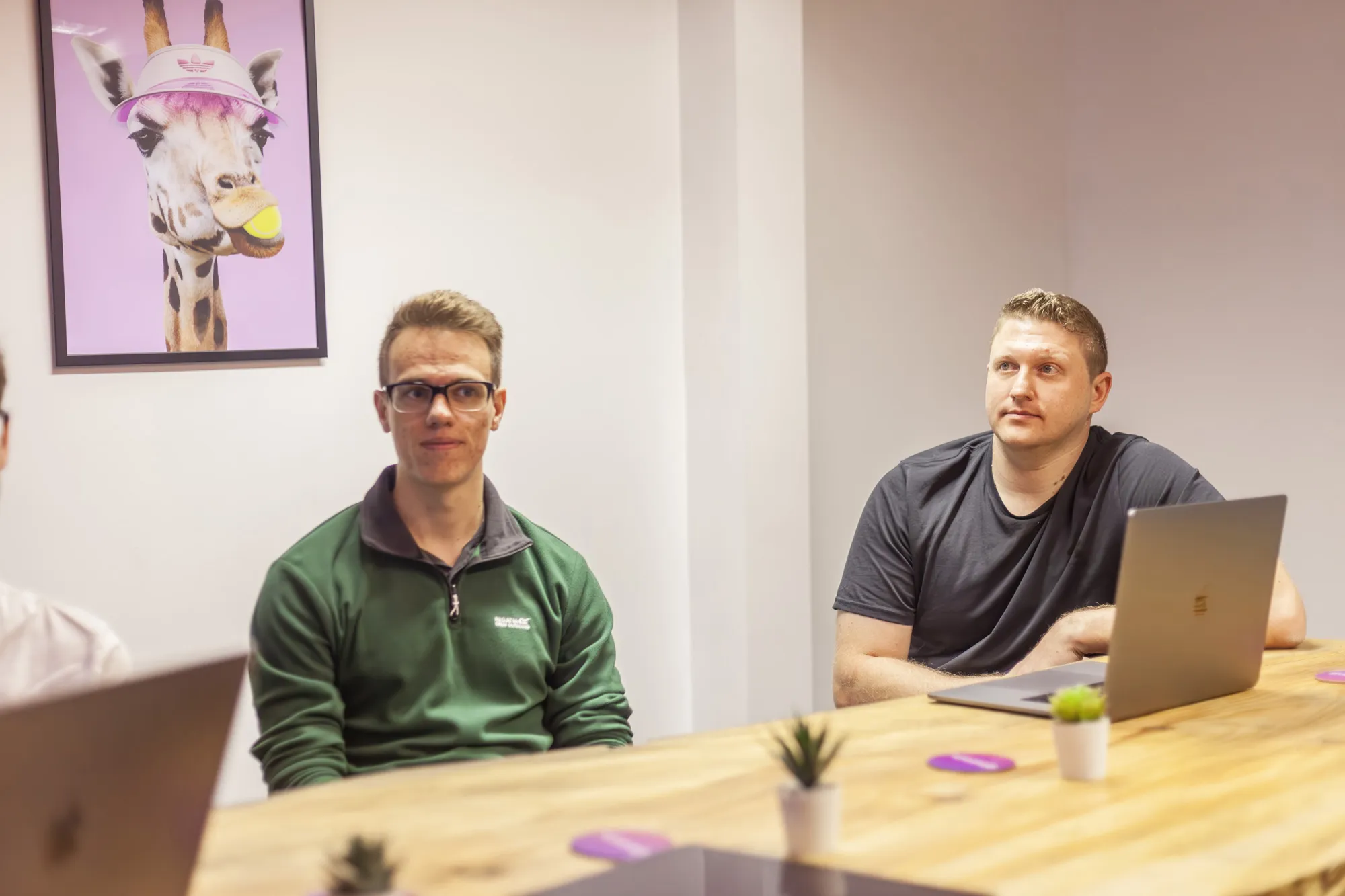 Two men sitting at a wooden table with laptops looking on