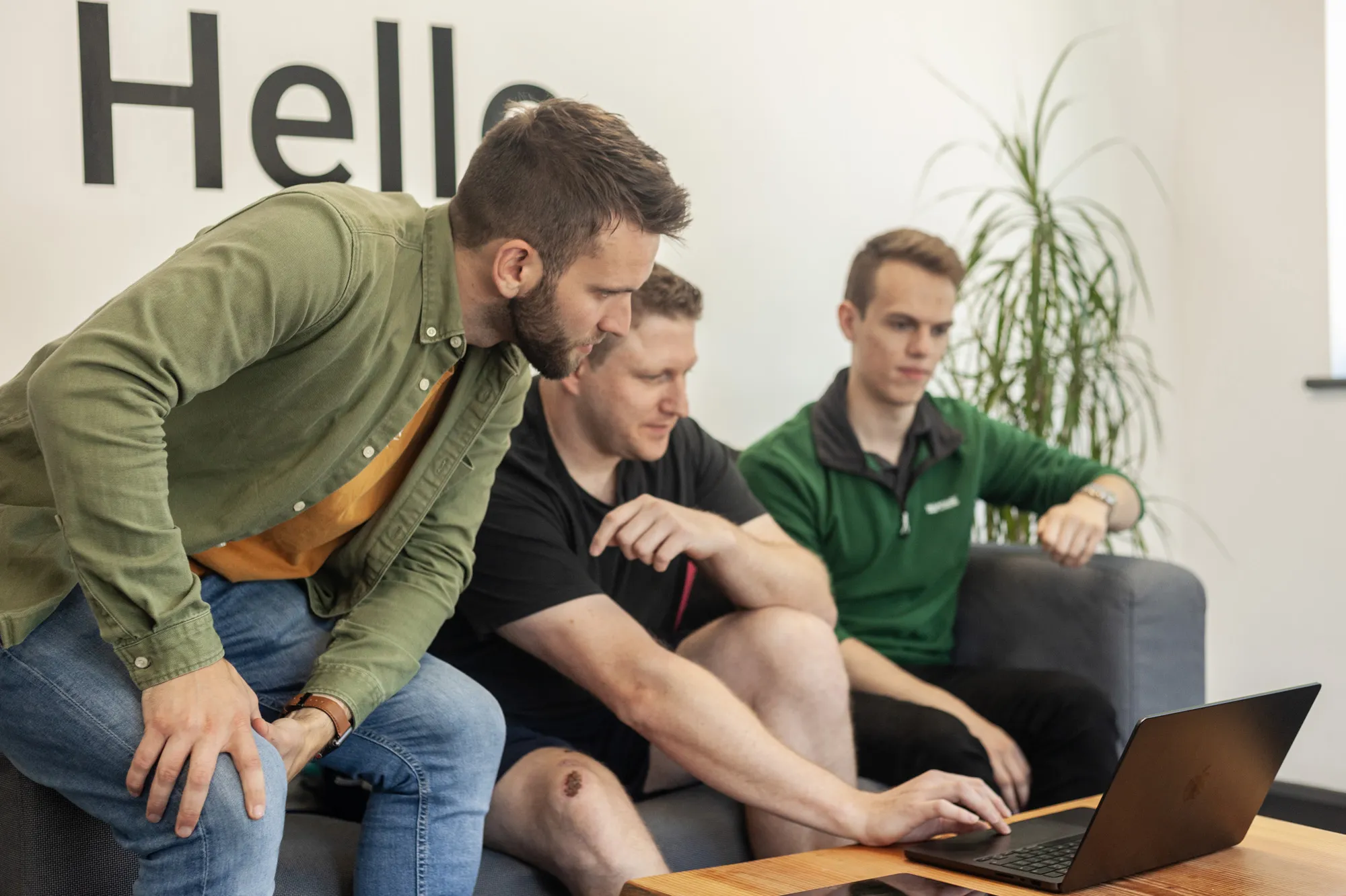 Three men sat on a grey sofa in a bright room looking at a laptop on a coffee table
