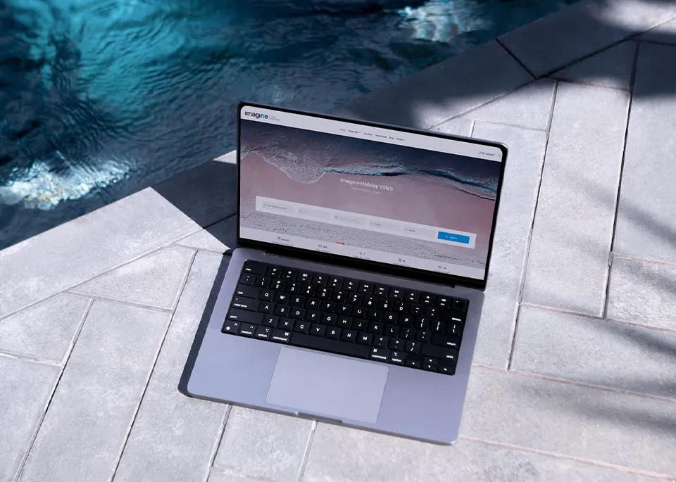 A MacBook on poolside tiles displaying the Imagine Villas website homepage