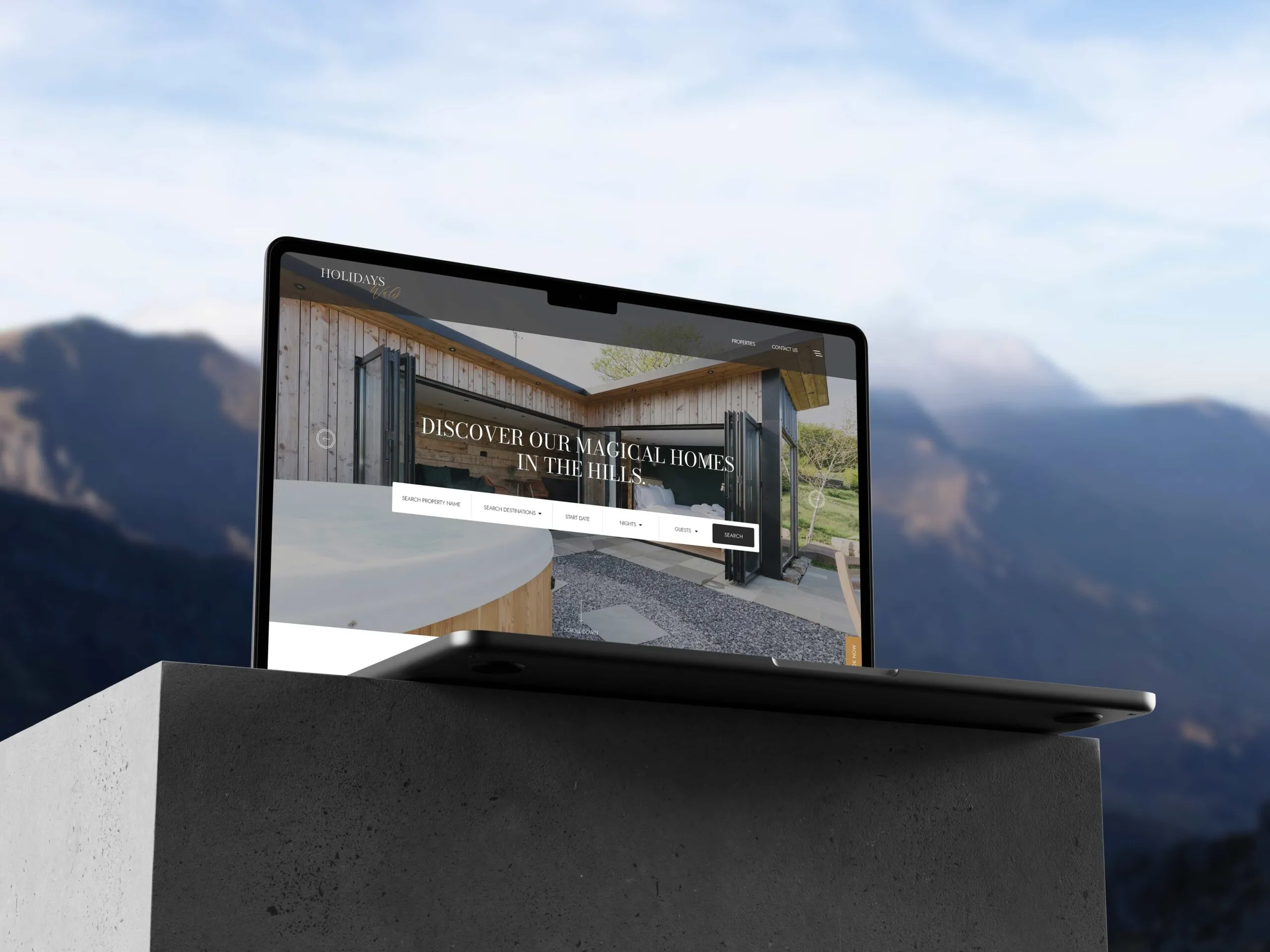 A MacBook displaying a the Holidays Wales homepage on a concrete platform with a blurred mountainous background