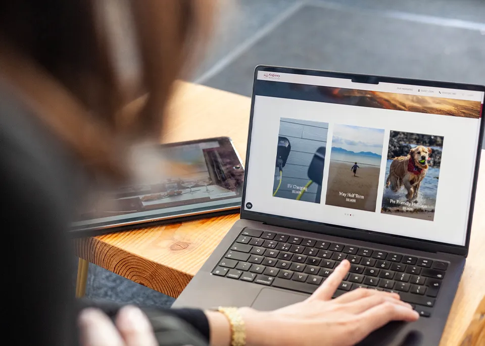 A person using a MacBook on a wooden table displaying a section of the Anglesey Holiday Lettings website with an iPad next to it