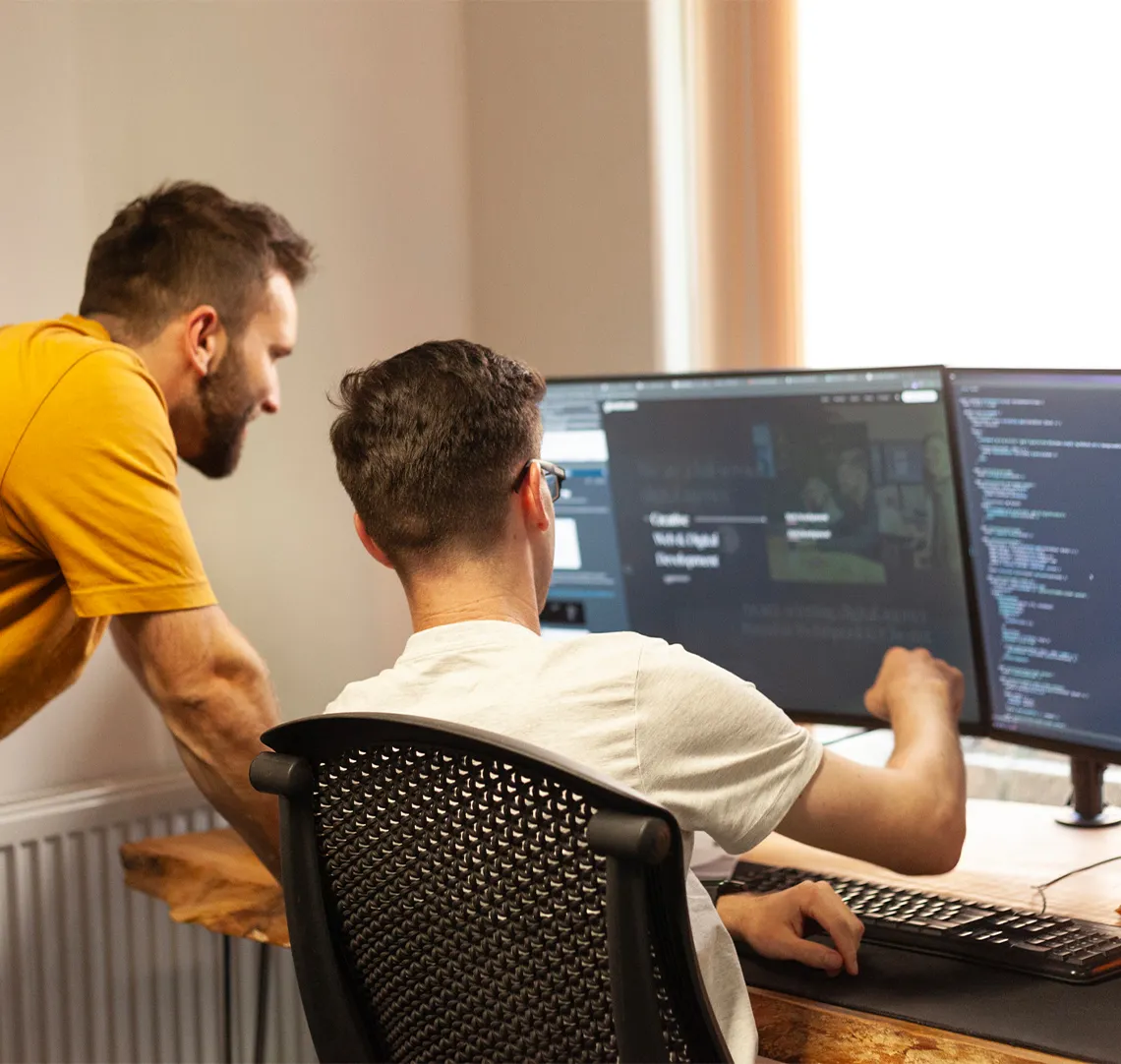 Two people collaborate at a desk with multiple monitors displaying code.